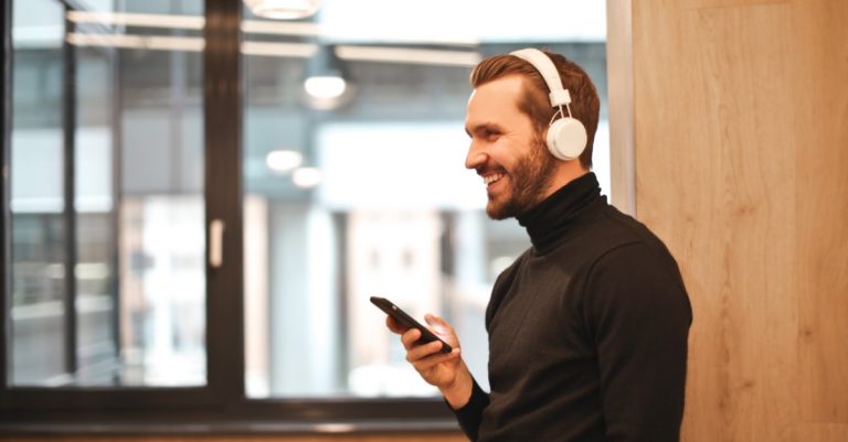A man wearing headphones smiles while holding his mobile phone.