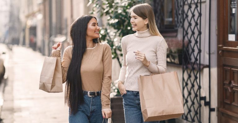 Two girls walk down a high street carrying shopping bags.
