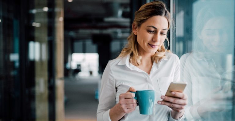 A woman smiles as she looks down at an SMS on her phone. She is in business attire and is holding a mug of coffee.