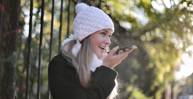 A woman speaks to a voice bot on her mobile phone. She is wearing a white beanie.